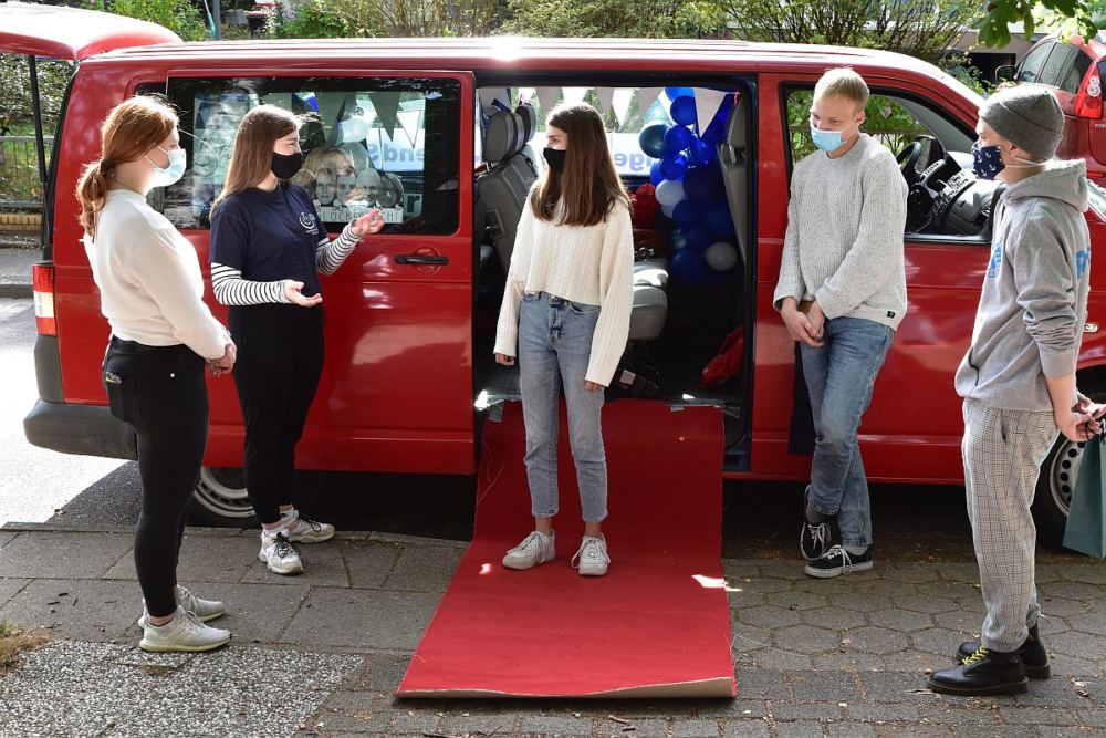 4 Teamer segnen eine Newcomerin auf einem roten Teppich vor einem Kleinbus.